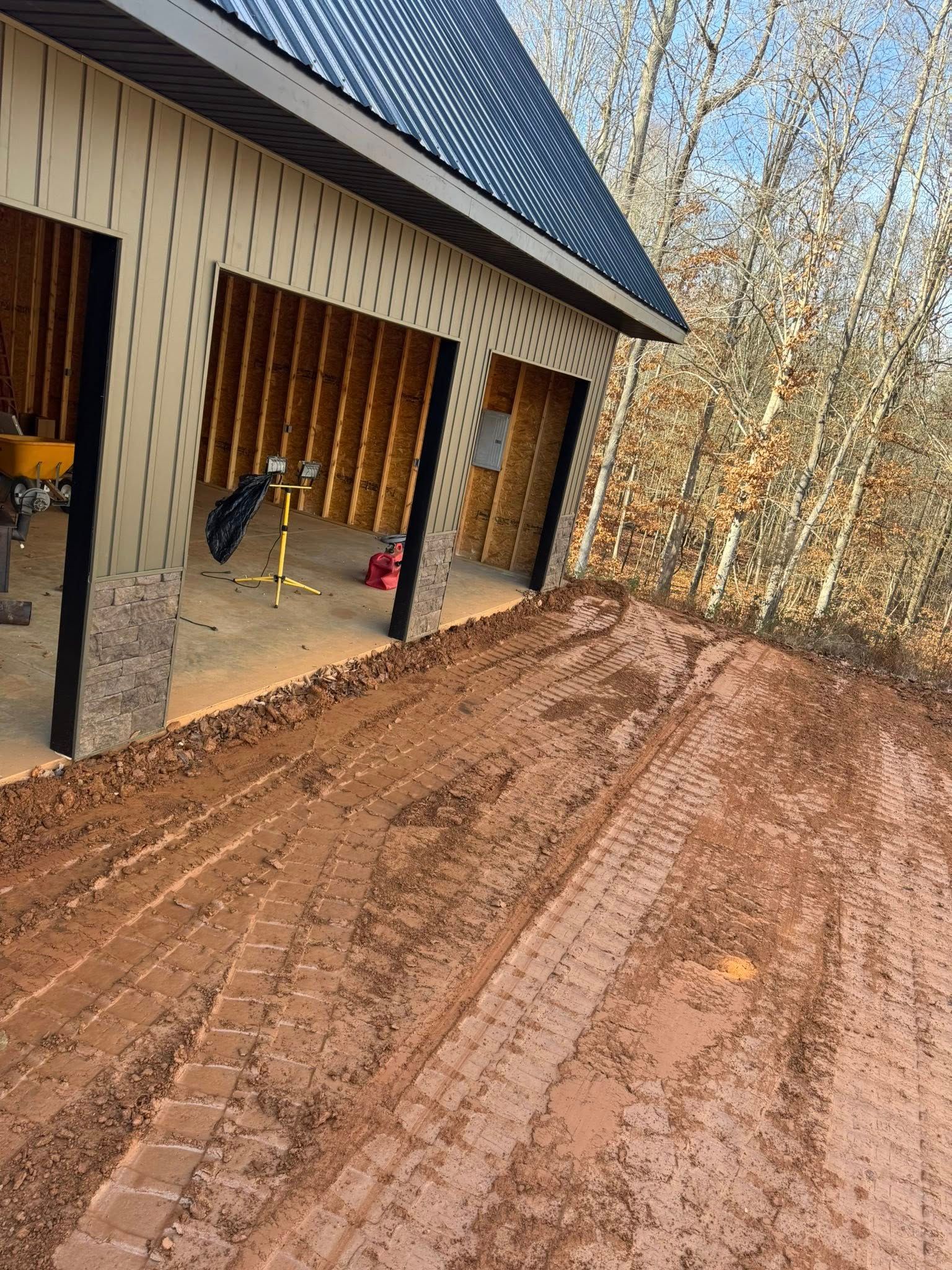 Red dirt driveway beside a house under construction with open garage bays and trees in the background