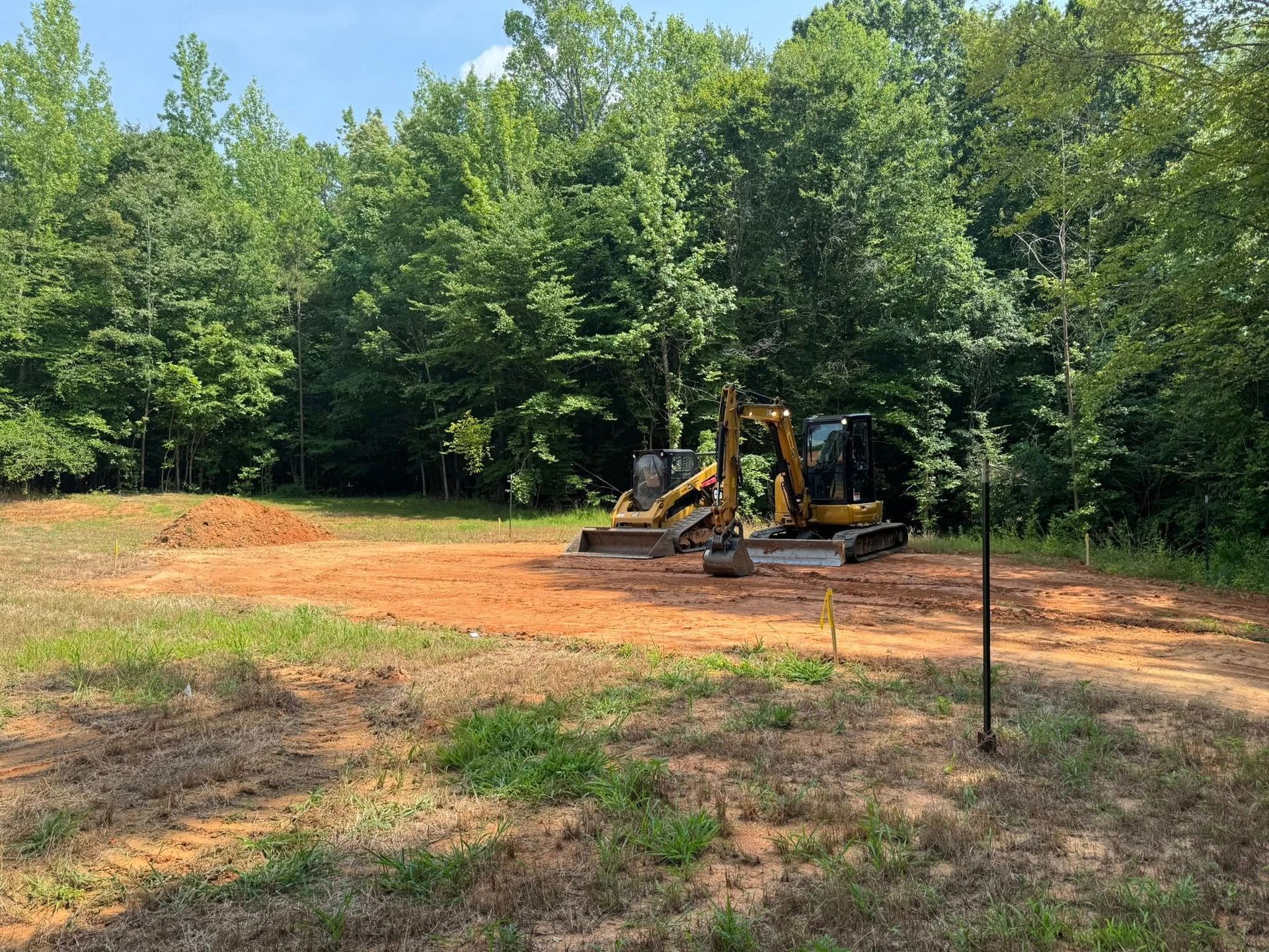 Yellow backhoe in a cleared dirt lot beside a wooded tree line under a sunny sky