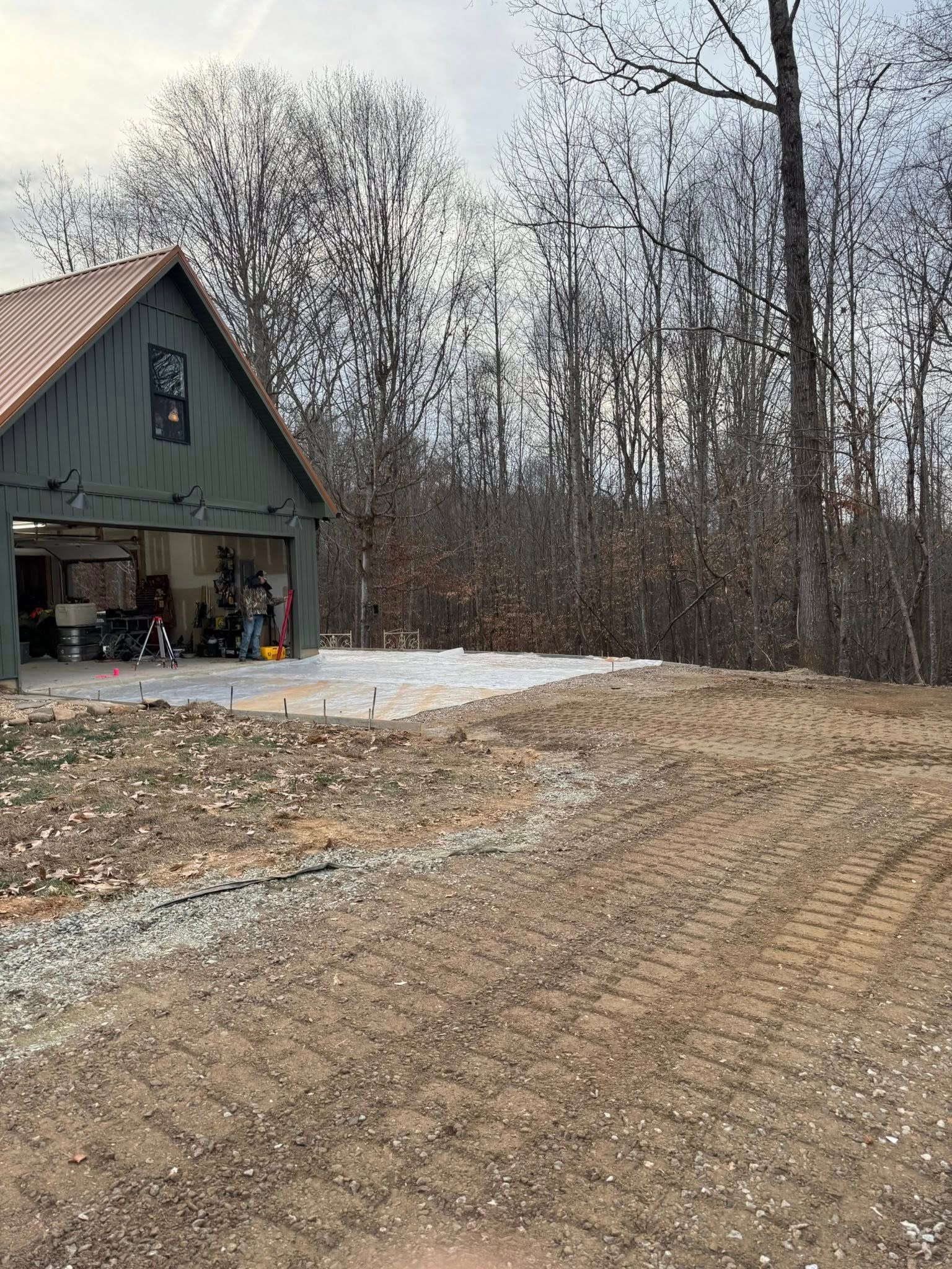 Rural driveway beside a green garage and wooded yard, with snow patches and bare trees.