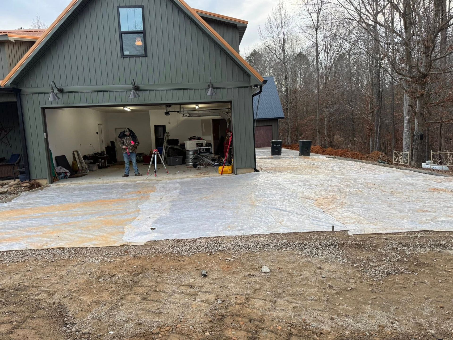 Green barn with open garage doors beside a gravel driveway and wooded yard; two people stand inside.