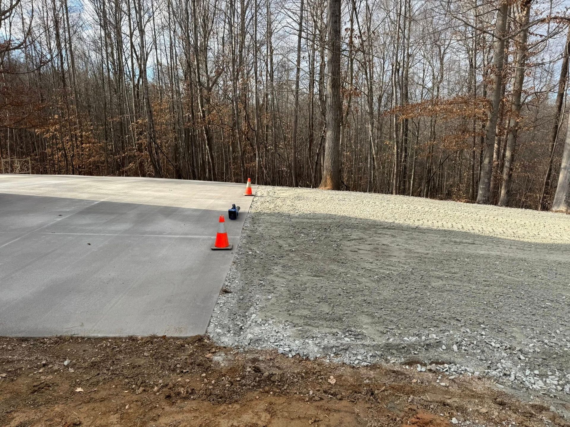 Orange cones marking the edge of a paved road beside a gravel shoulder in a wooded area