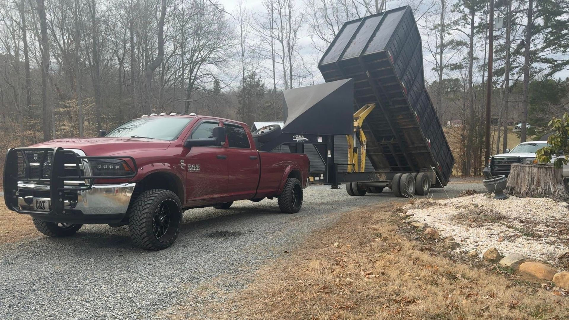 Red pickup truck beside a raised black dump bed on a gravel driveway in a wooded yard.