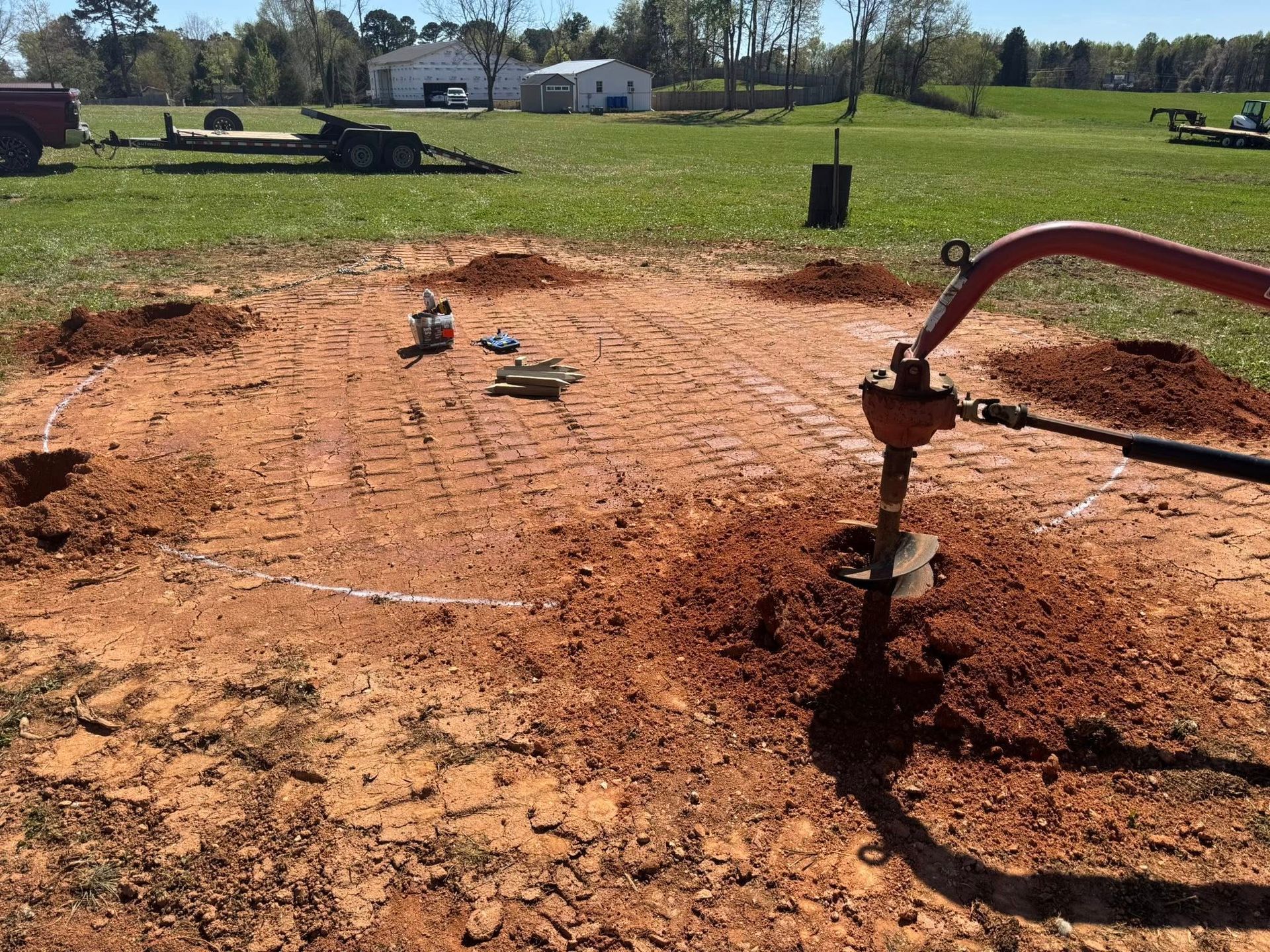Red dirt construction site with an auger drilling a hole in the ground and tools scattered nearby.