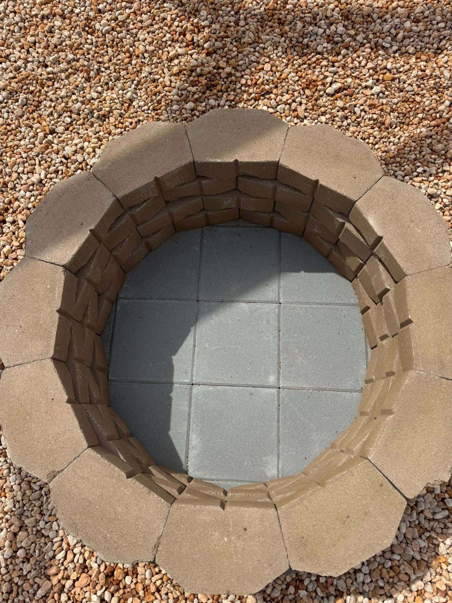 Top-down view of a circular stone-lined pit with gray floor tiles, surrounded by gravel.