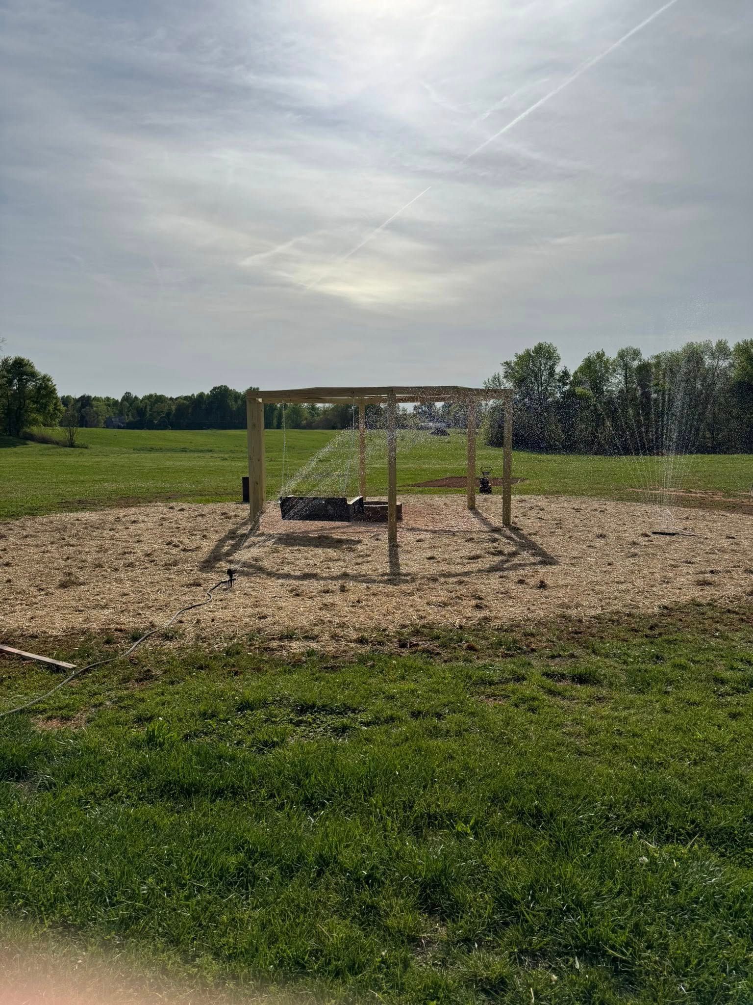 Small wooden playground frame on a woodchip field under a bright cloudy sky, with grassy fields behind it