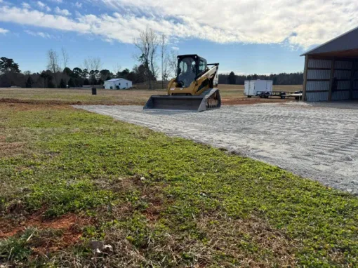 Yellow skid steer on gravel lot beside a barn, with grassy foreground and cloudy sky.