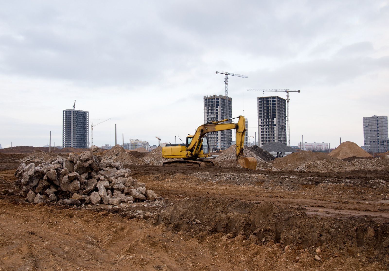 Yellow excavator at a muddy construction site with high-rise buildings in the background