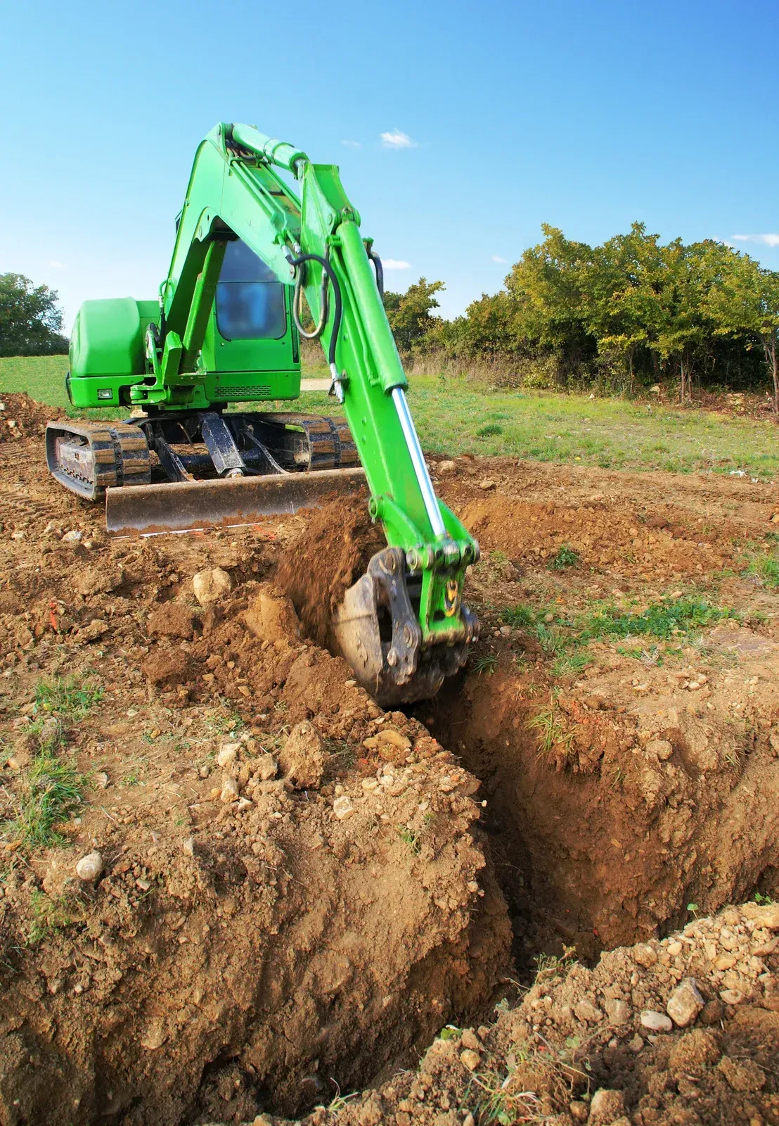 Green excavator digging a trench in a dirt field under a clear blue sky