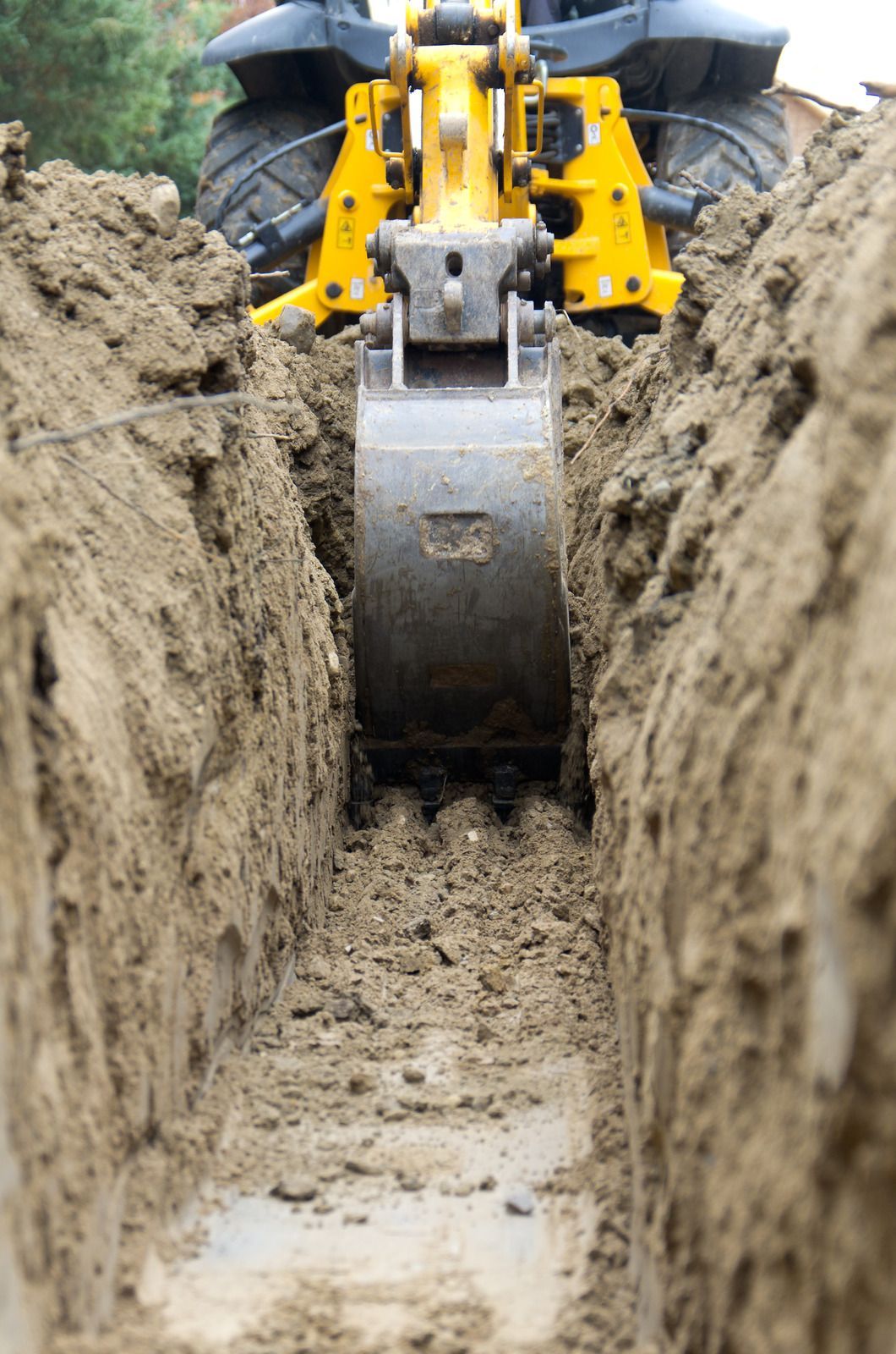Yellow excavator bucket digging a narrow trench in muddy soil