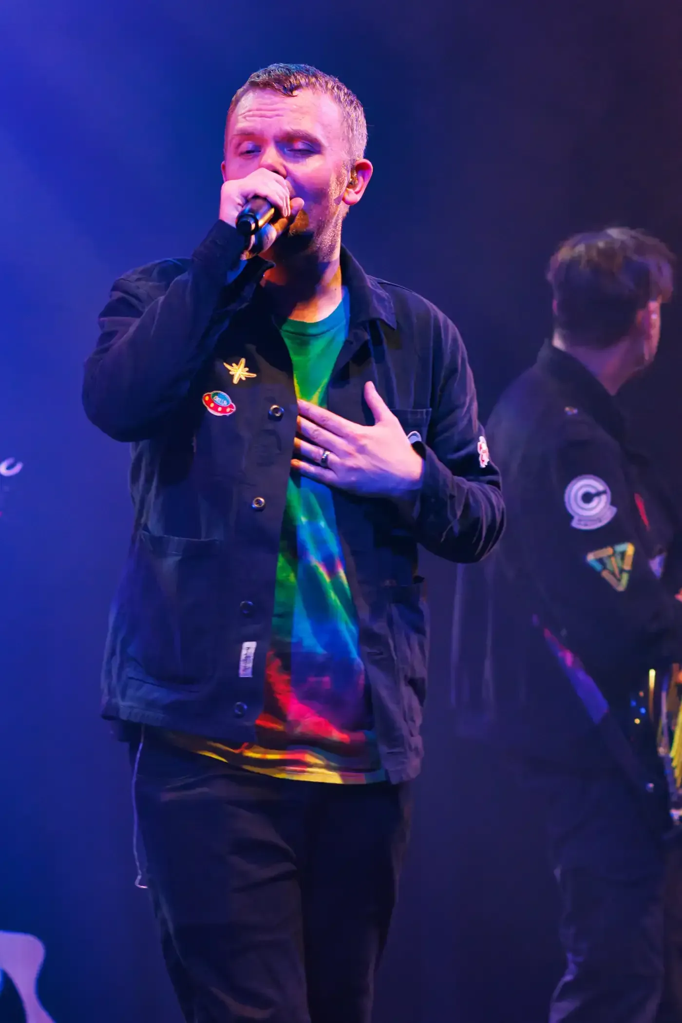 Singer holding a microphone on stage under purple lighting, wearing a black jacket and colorful shirt