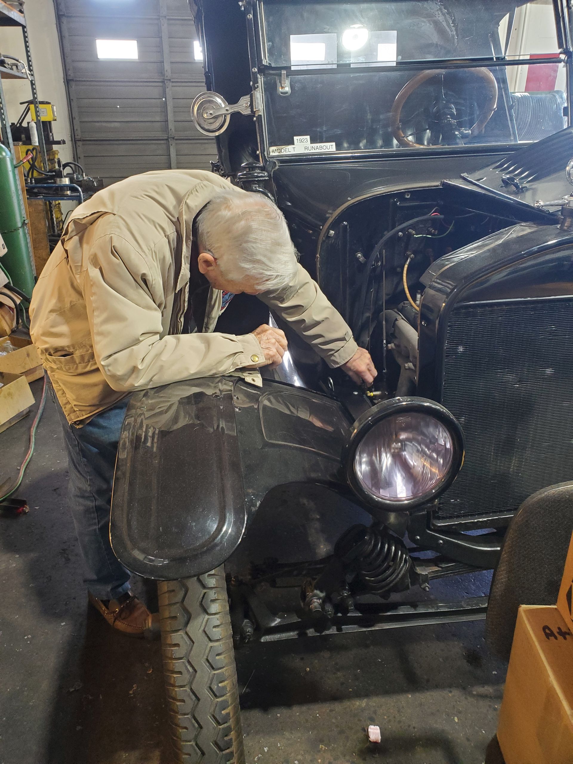 Man working on a black vintage car in a garage, wearing a tan jacket, inspecting the engine. | Hal's Auto Care