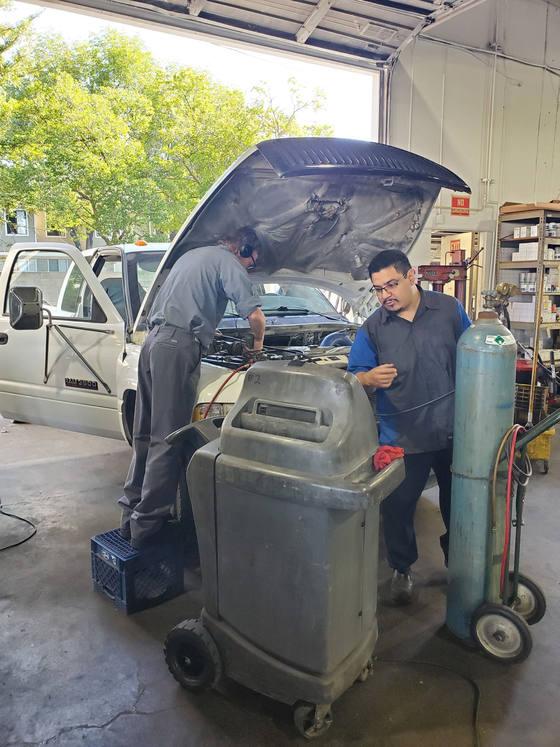 Two mechanics working on a truck in a shop. One under the hood, the other near a machine and a blue gas tank. | Hal's Auto Care