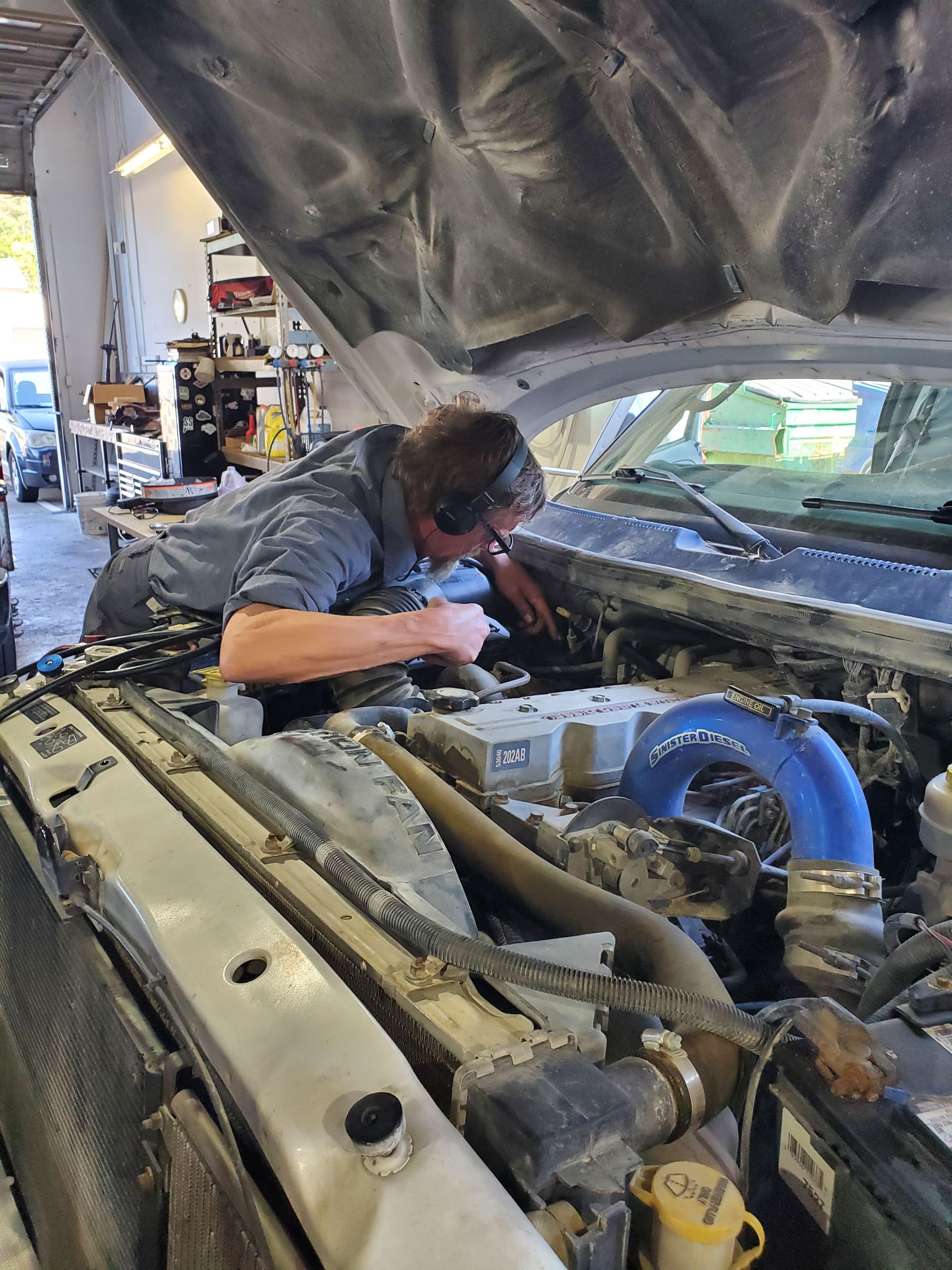 Mechanic working on a truck engine; under the open hood. Wearing ear protection. Gray and blue. | Hal's Auto Care