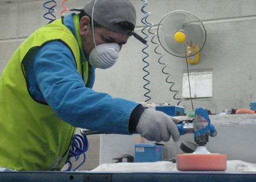 Person in safety gear using a polishing tool on a surface, indoors, near a fan.