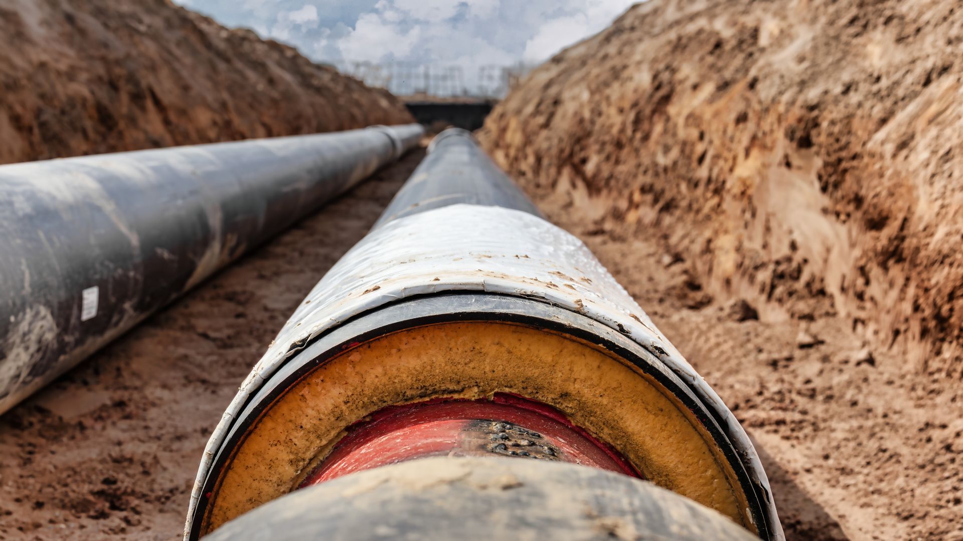Large pipes with insulation installed in a trench. Brown earth visible; cloudy sky overhead.