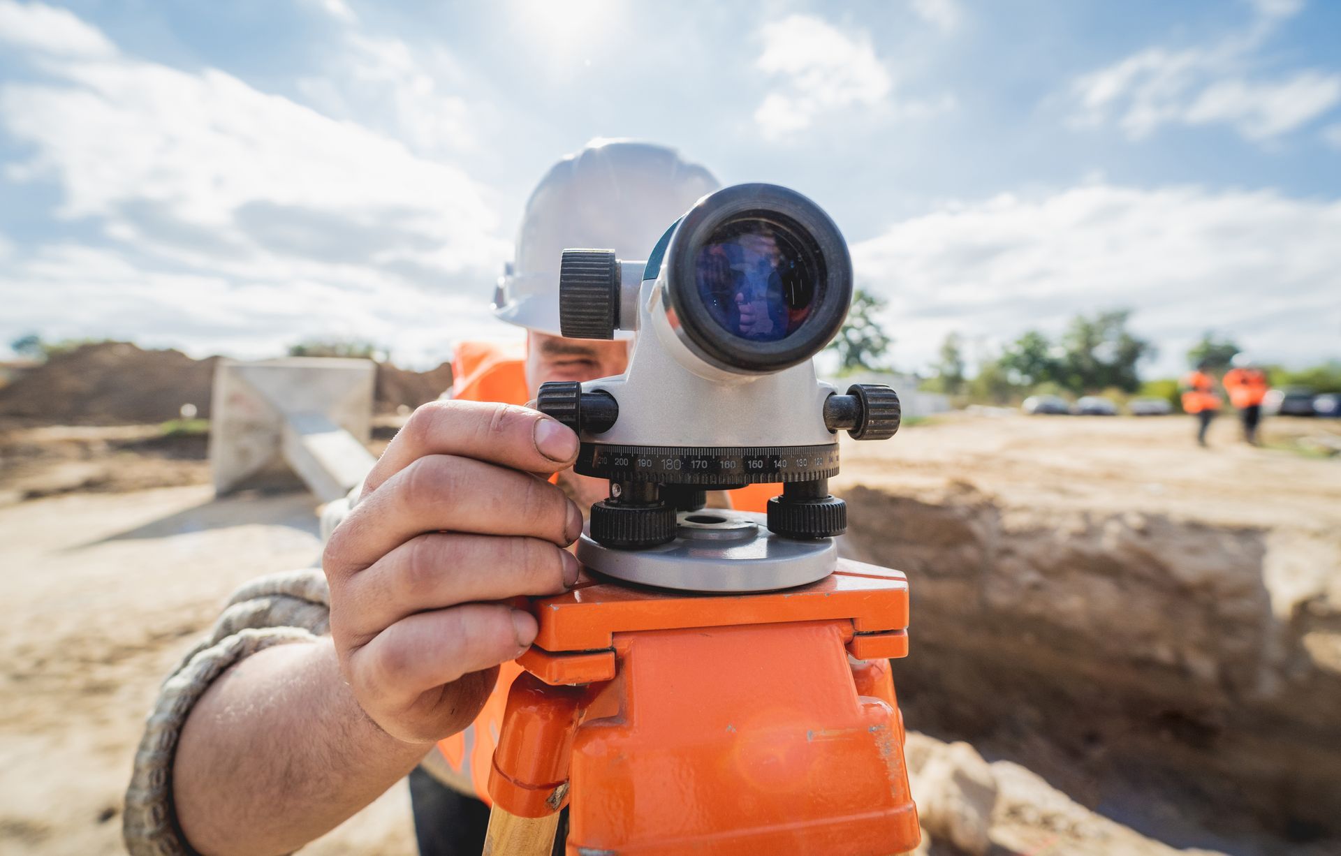 Person using a surveying level outdoors, focusing on a construction site. Bright, sunny day.