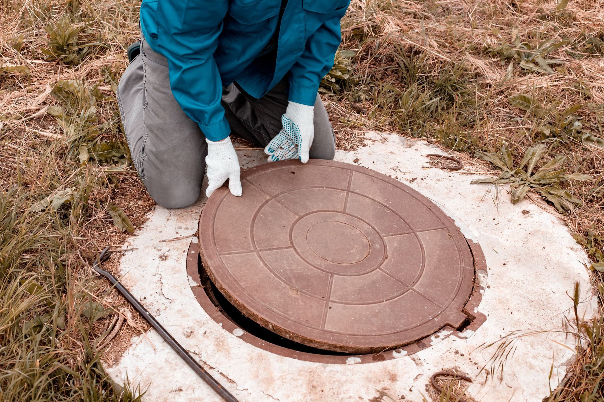 Person opening a septic tank cover in a grassy area, wearing gloves and teal shirt.