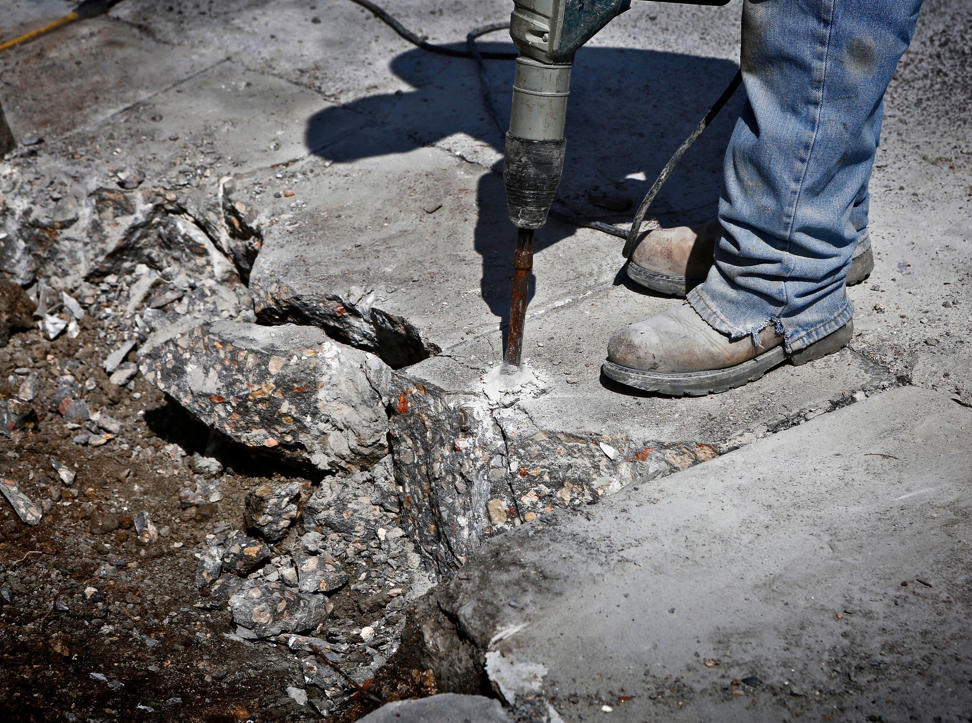A worker in jeans and work boots uses a jackhammer on concrete.