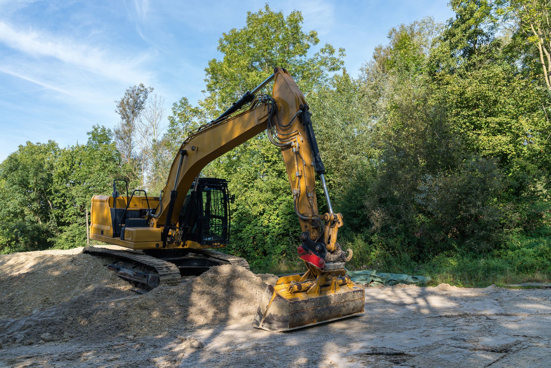 Yellow excavator digging into a pile of dirt, with trees in the background under a blue sky.