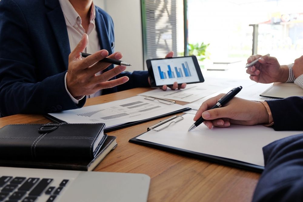 Pessoas de terno em uma mesa, discutindo gráficos de negócios em um tablet e documentos; ambiente de escritório.