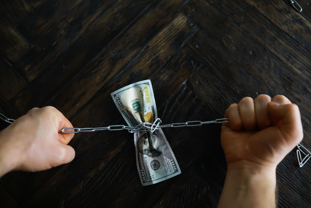 Mãos puxando uma corrente que envolve um maço de notas de dólar americano sobre uma superfície de madeira escura.