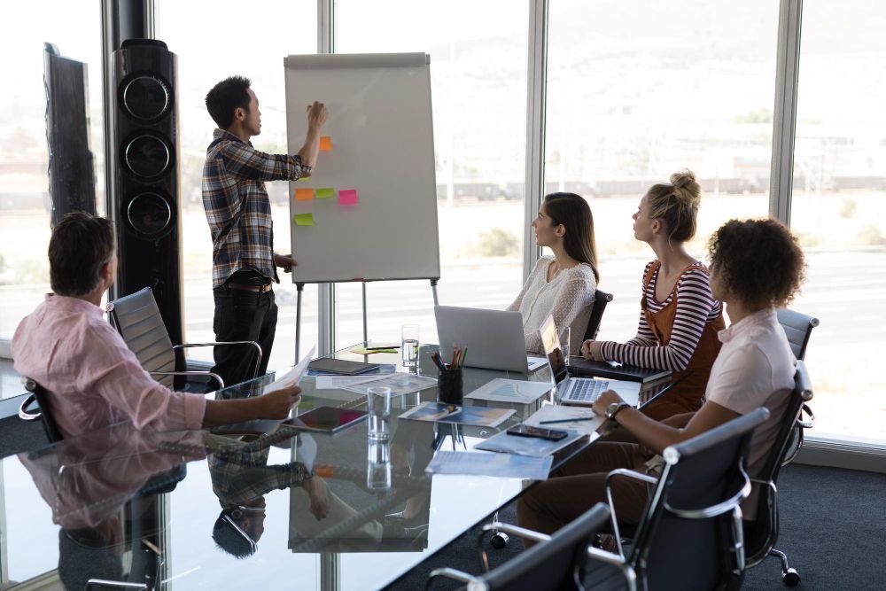 Um grupo diverso em uma reunião, com um homem apresentando em um quadro branco, post-its coloridos, perto de uma janela.
