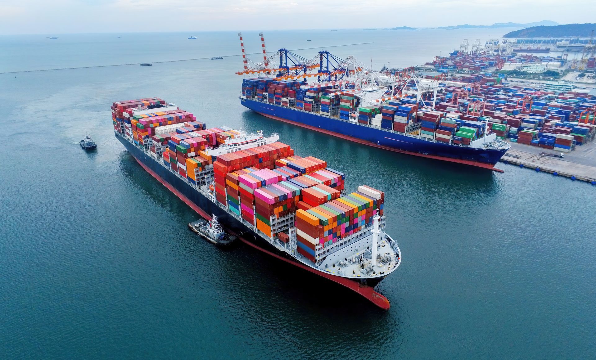Dois grandes navios de carga em um porto, carregados com contêineres coloridos. Guindastes e um porto ao fundo.
