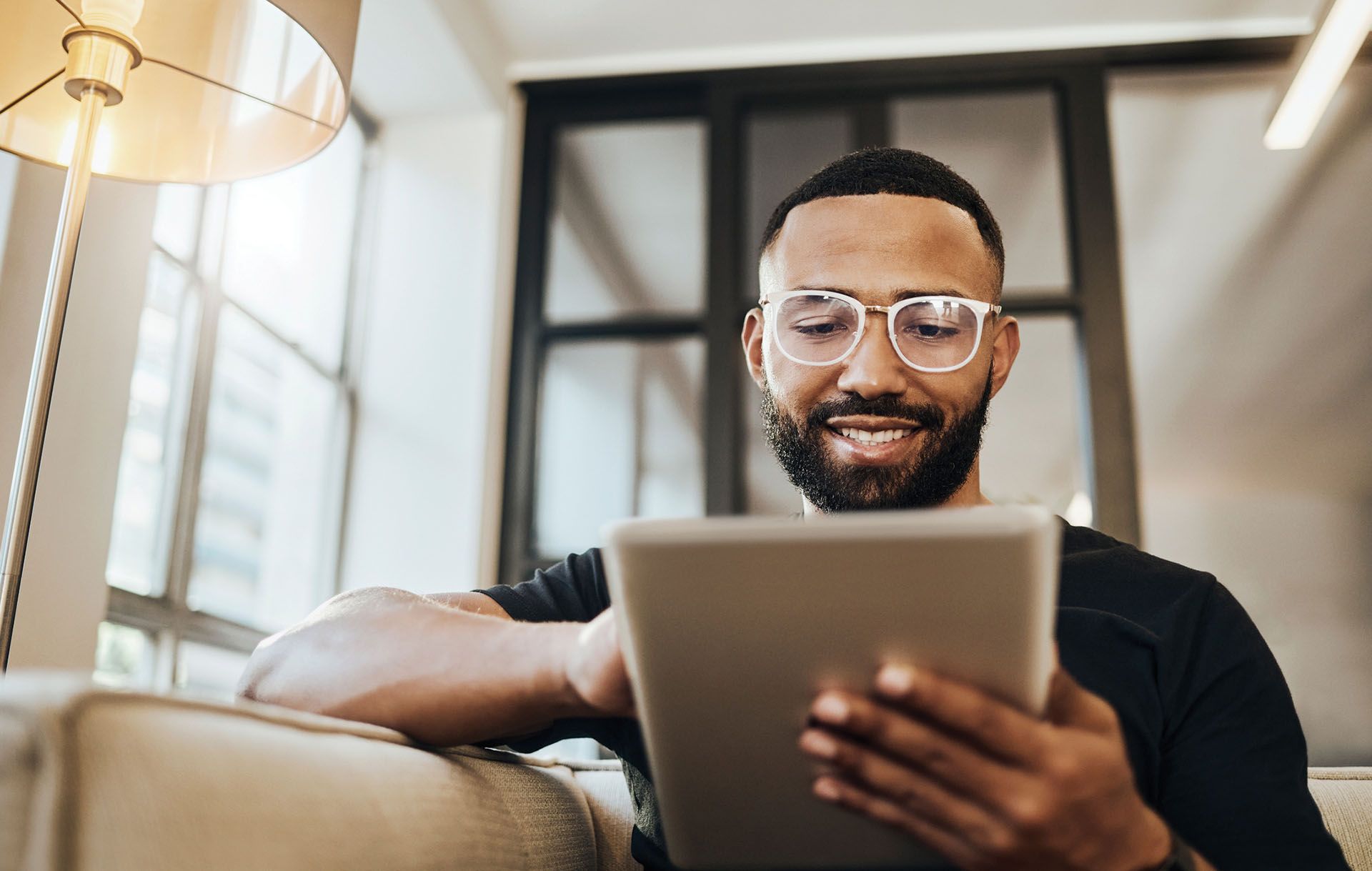 Man with glasses smiles while looking at tablet on a couch.