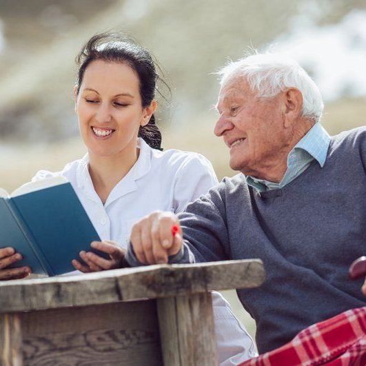 A caregiver reads from a blue book to a person sitting outdoors at a wooden table.