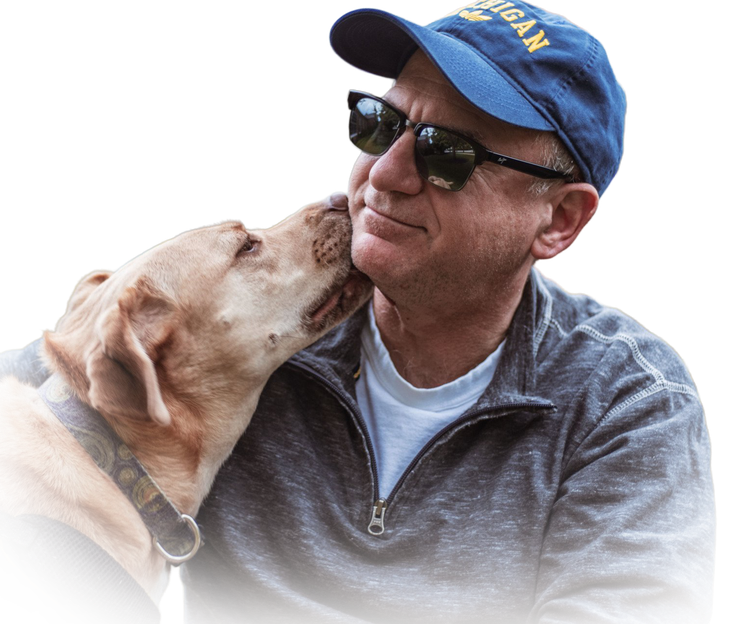 A person wearing a blue baseball cap and sunglasses is being affectionately licked on the cheek by a light-colored dog.