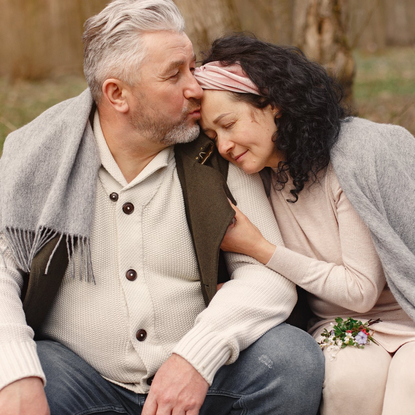 A couple wrapped in a grey blanket sitting in a forest setting, with the person on the left kissing the other's forehead.