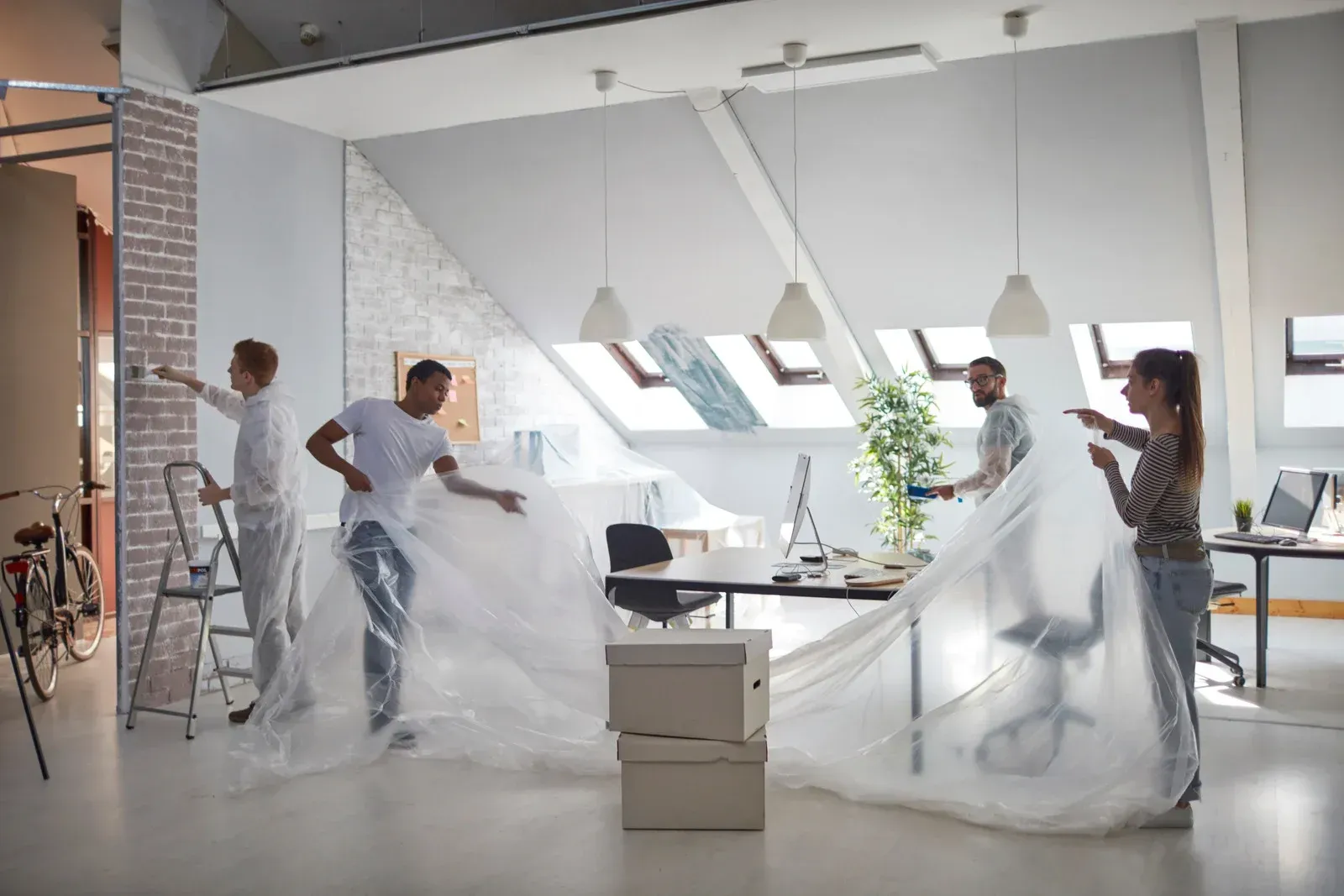 People covering furniture and painting in an office with skylights, preparing for renovation.