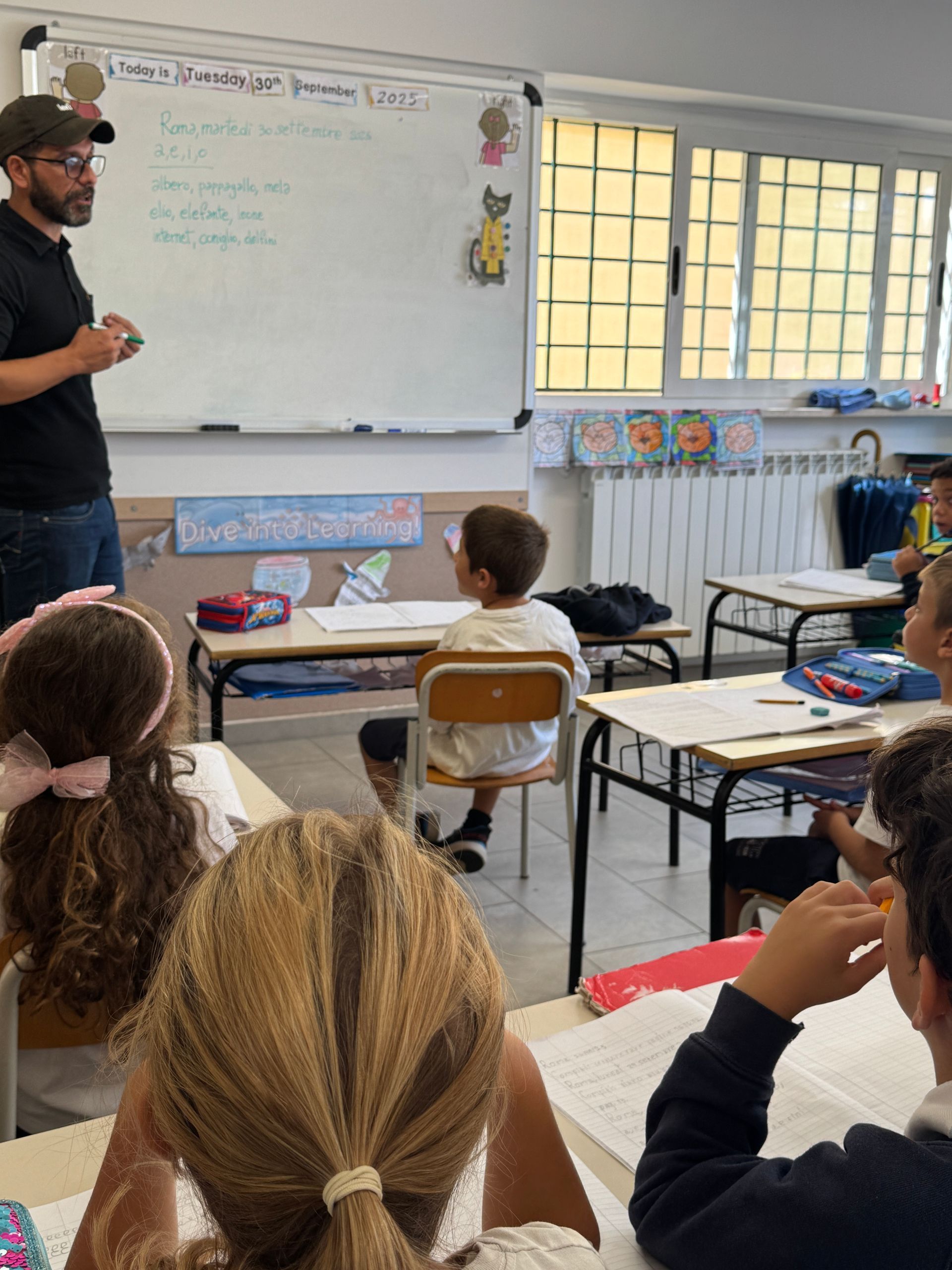 L'insegnante in classe scrive alla lavagna. Gli studenti sono seduti ai banchi e guardano l'insegnante.