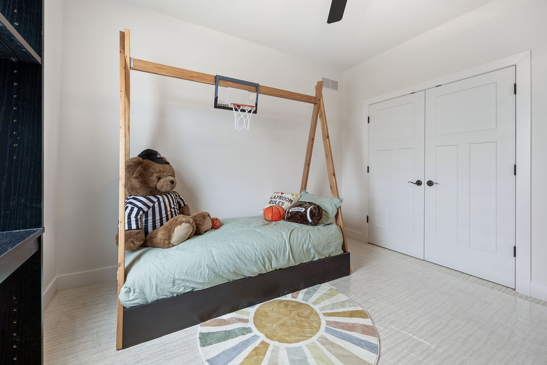 A bedroom with a teddy bear sitting on a bed and a basketball hoop hanging from the ceiling.