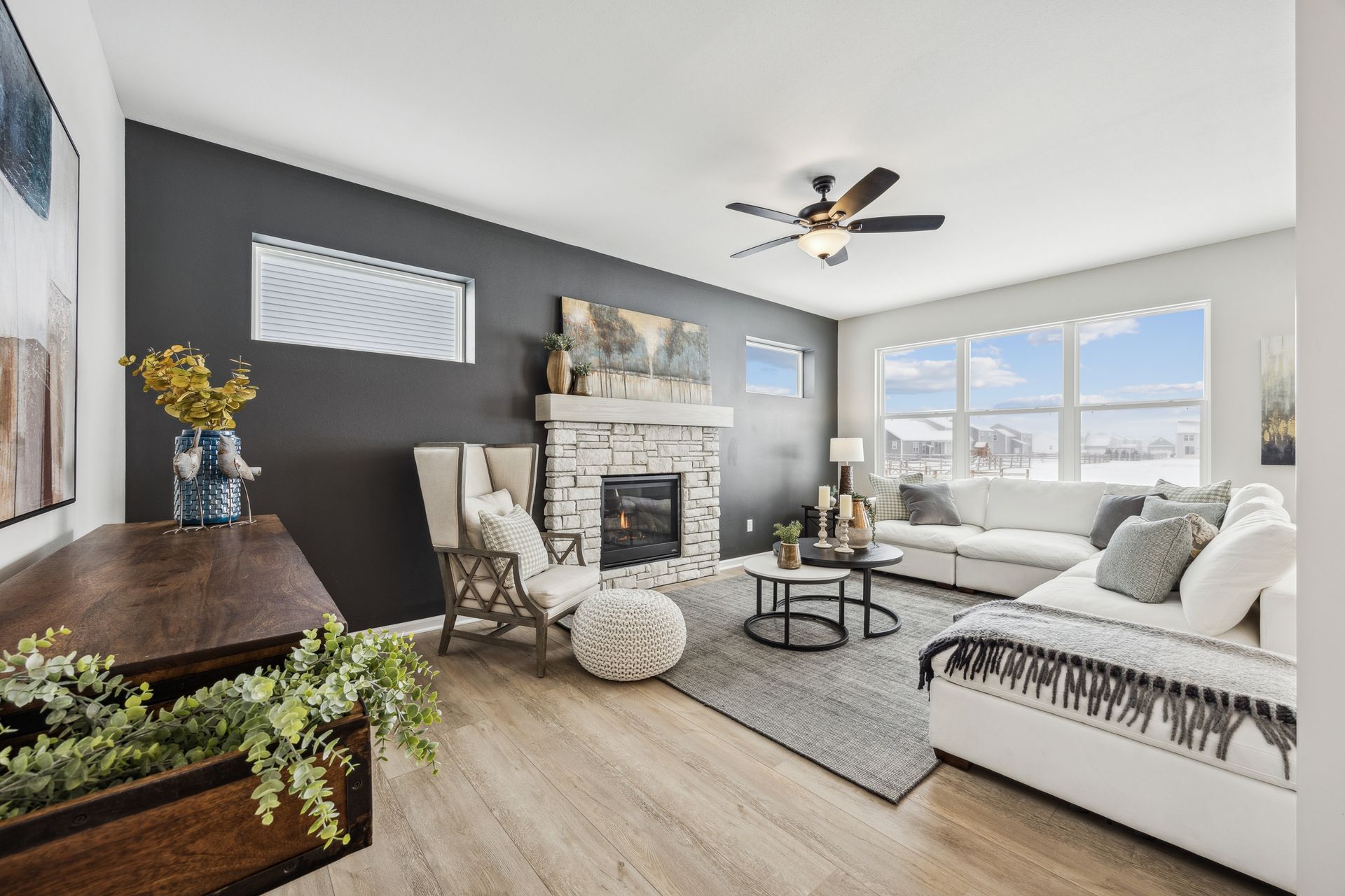 Living room with dark gray accent wall, stone fireplace, white sofa, and large windows with a winter view.