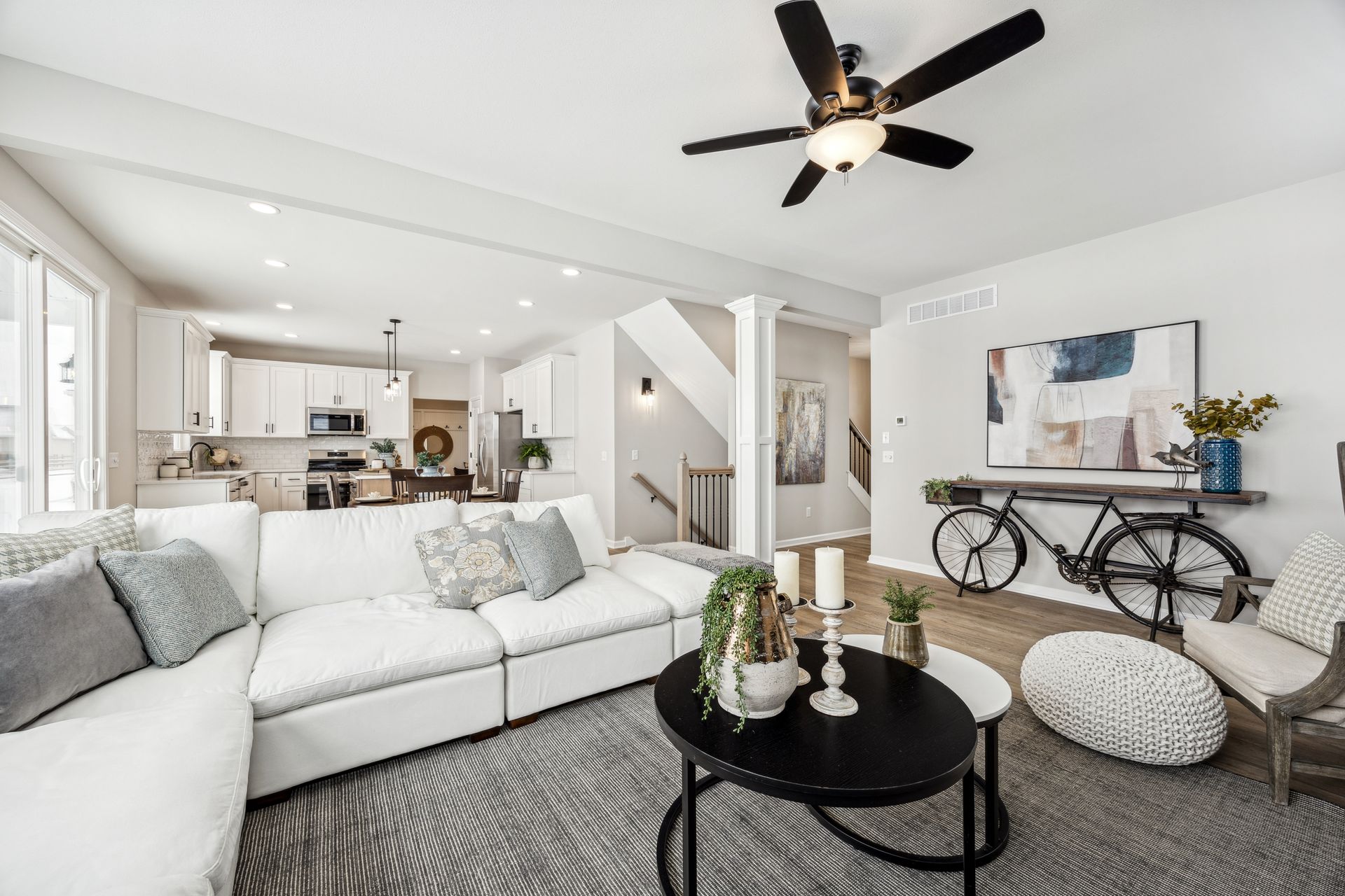 Modern living room with white sectional sofa, black coffee table, bicycle wall art, and open kitchen.