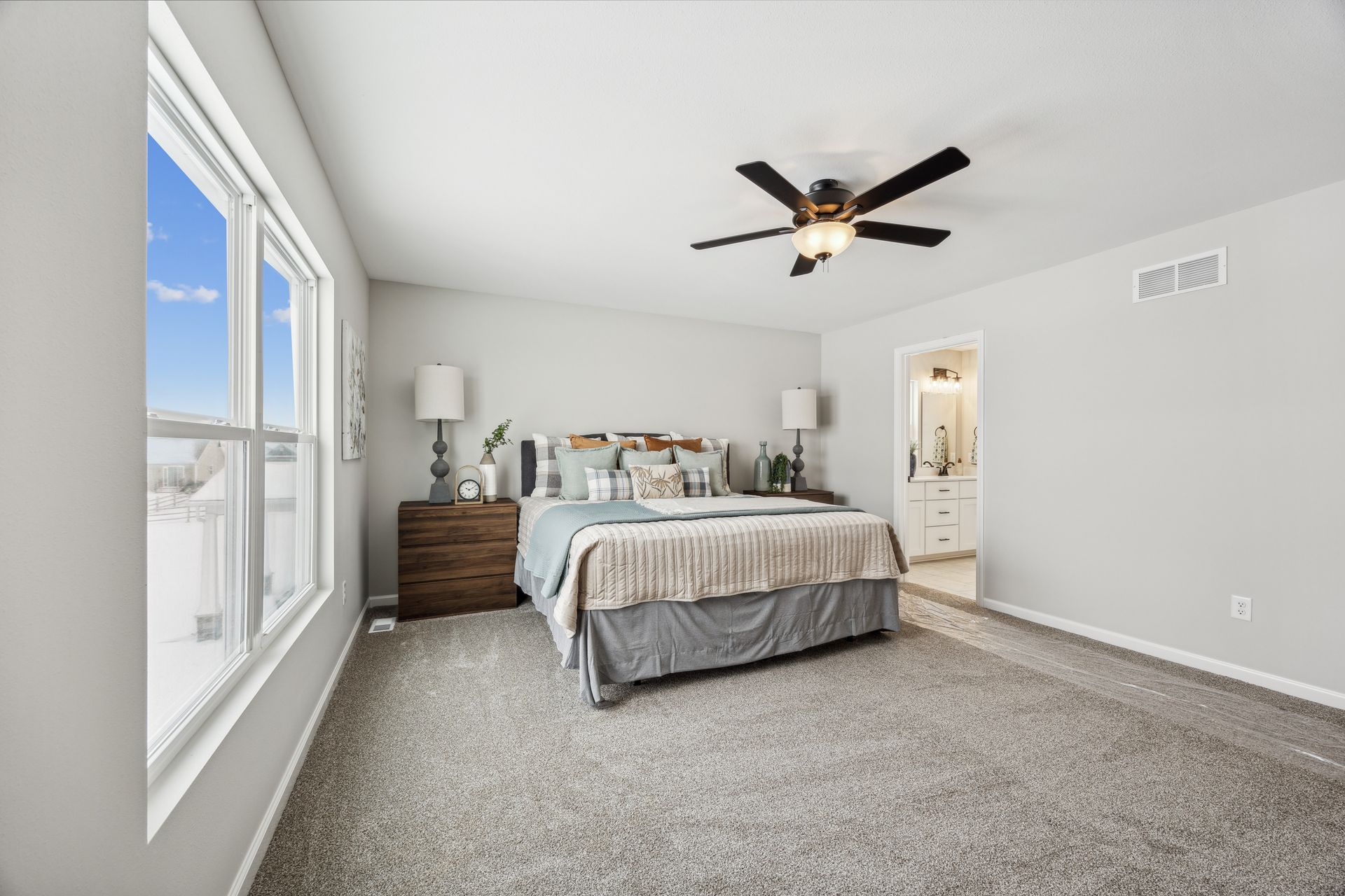 Bedroom with large window, bed, and ceiling fan. Neutral colors, carpeted floor. Doorway to bathroom.