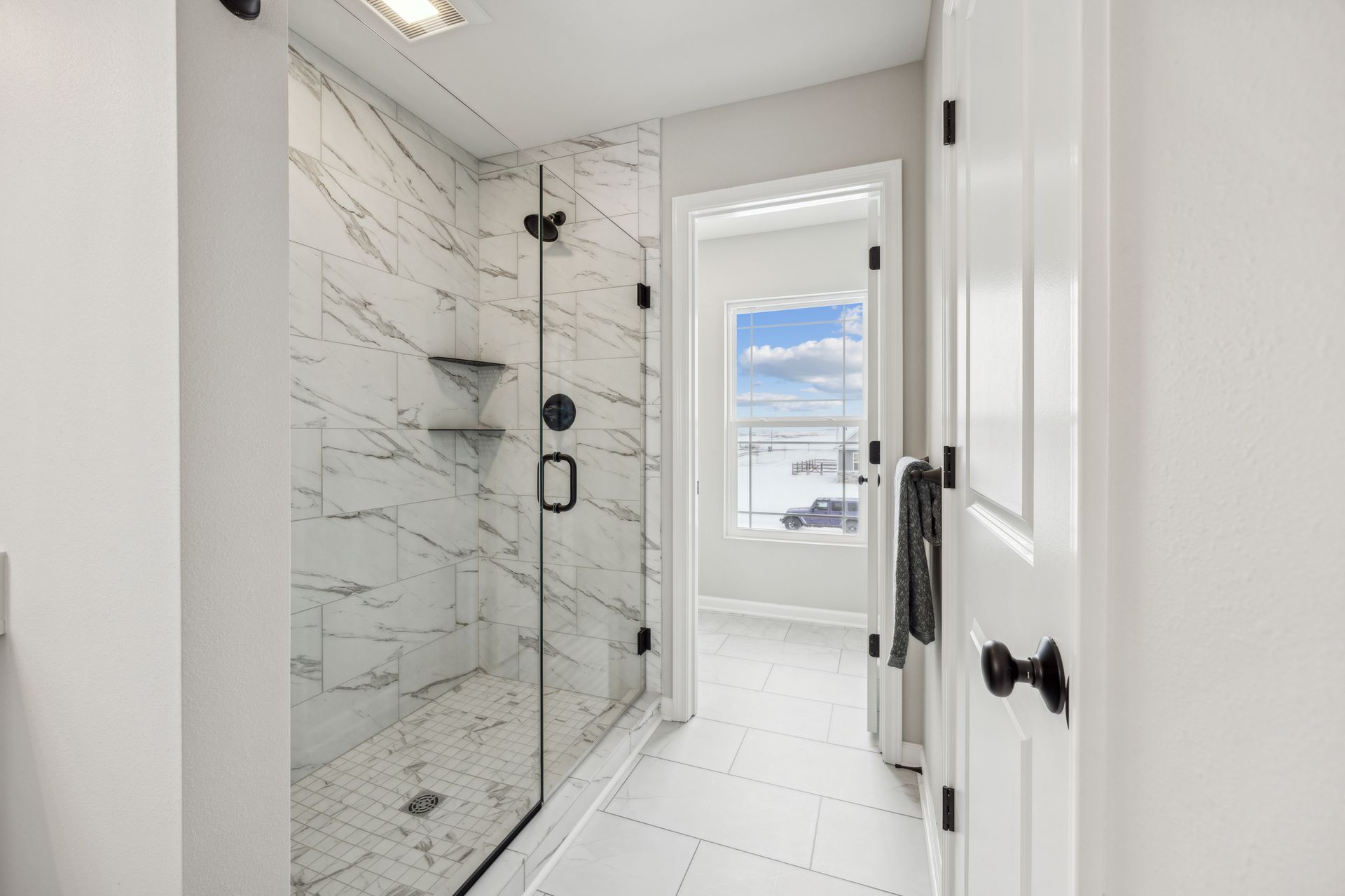 Modern white bathroom with marble shower, glass door, and hallway leading to a window.