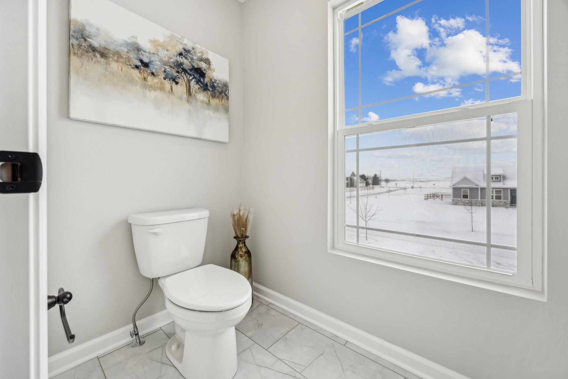 Toilet in a small bathroom with a snowy outdoor view through the window; artwork above the toilet.