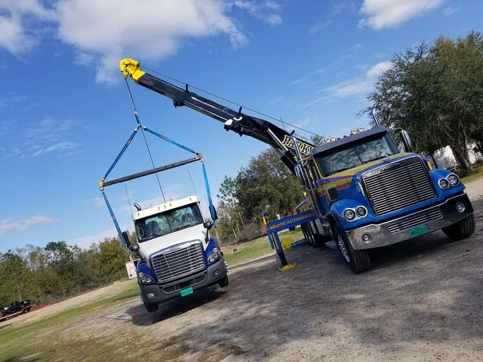 Two blue tow trucks lifting a white truck with a crane on a grassy roadside under a blue sky.