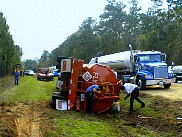 Truck overturned in a muddy ditch as people inspect it on a rural roadside