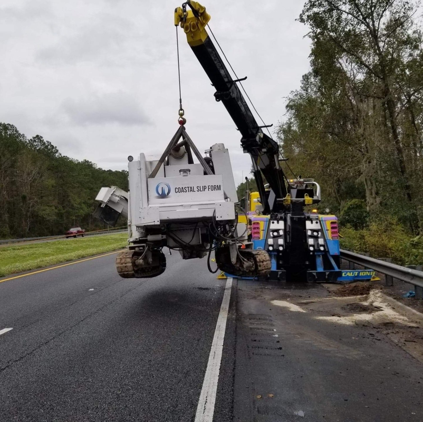 Tow truck lifting a white truck off a highway shoulder near a guardrail and trees