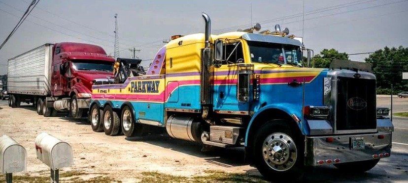 A large yellow, blue, and pink tow truck is towing a red semi-truck on a dirt road under a clear sky.