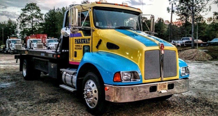 A large yellow, blue, and pink tow truck is towing a red semi-truck on a dirt road under a clear sky.