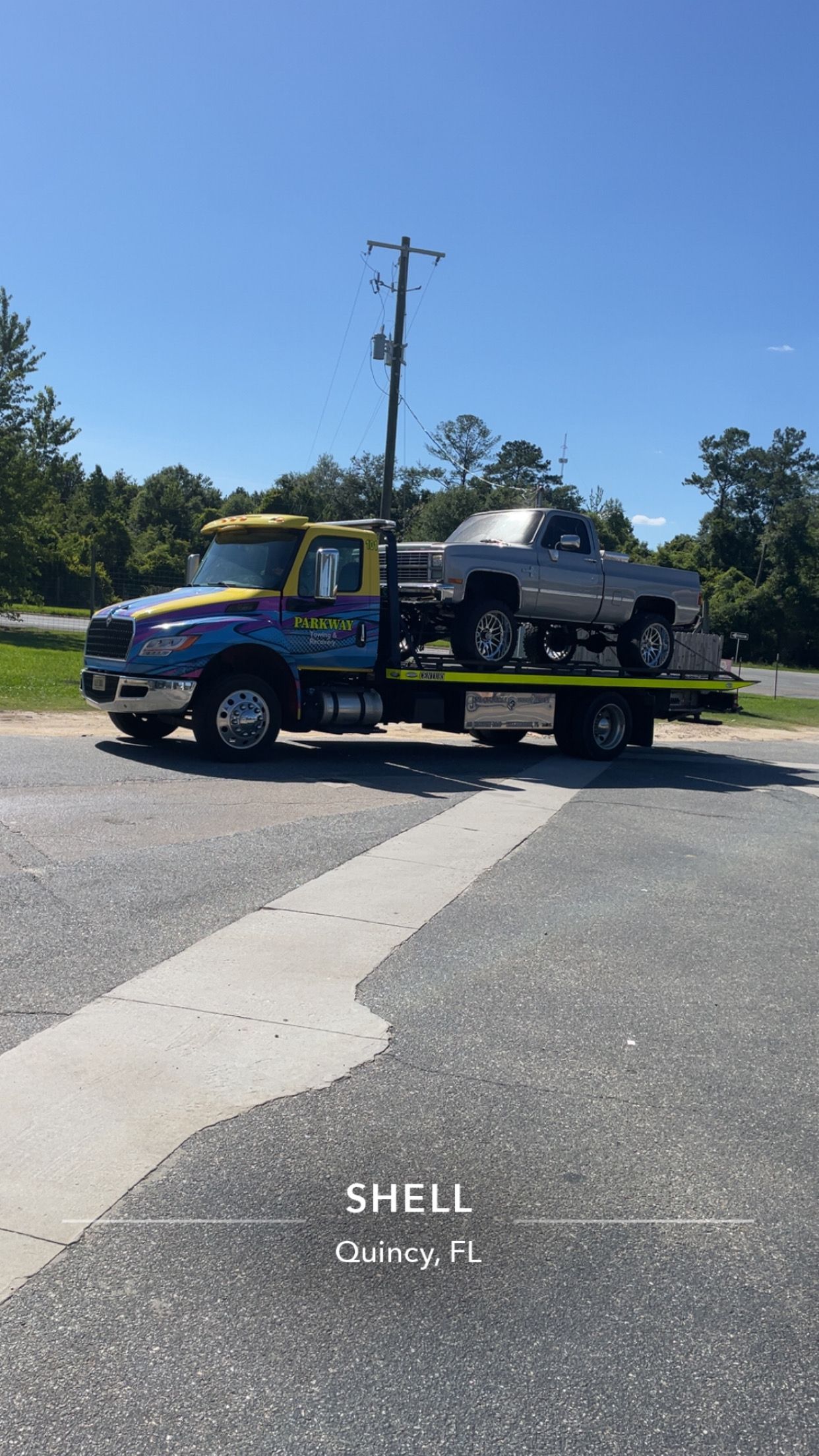 Blue tow truck carrying a black pickup on a flatbed in a Shell parking lot under a clear sky