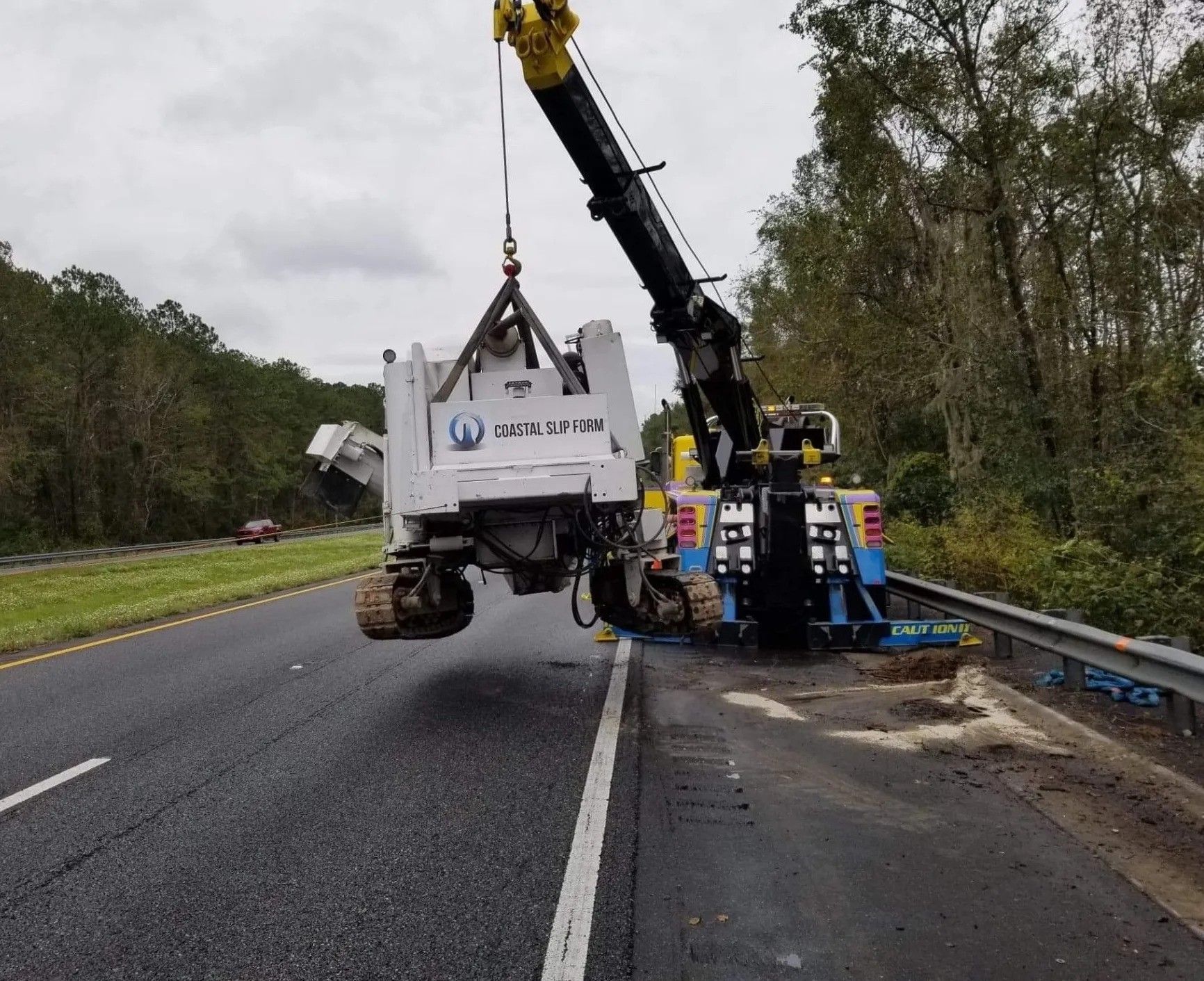 Crane lifting a white truck from a highway shoulder beside a guardrail in a wooded area