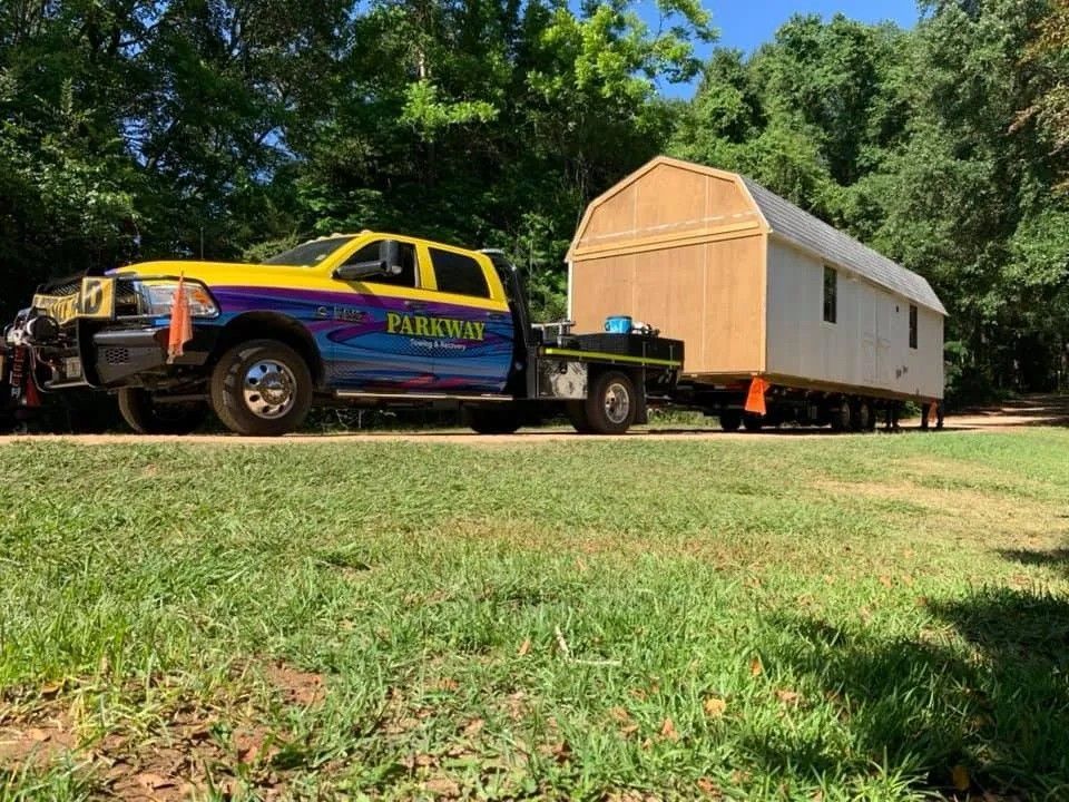 Blue-and-yellow pickup truck towing a white shed across a grassy yard under trees