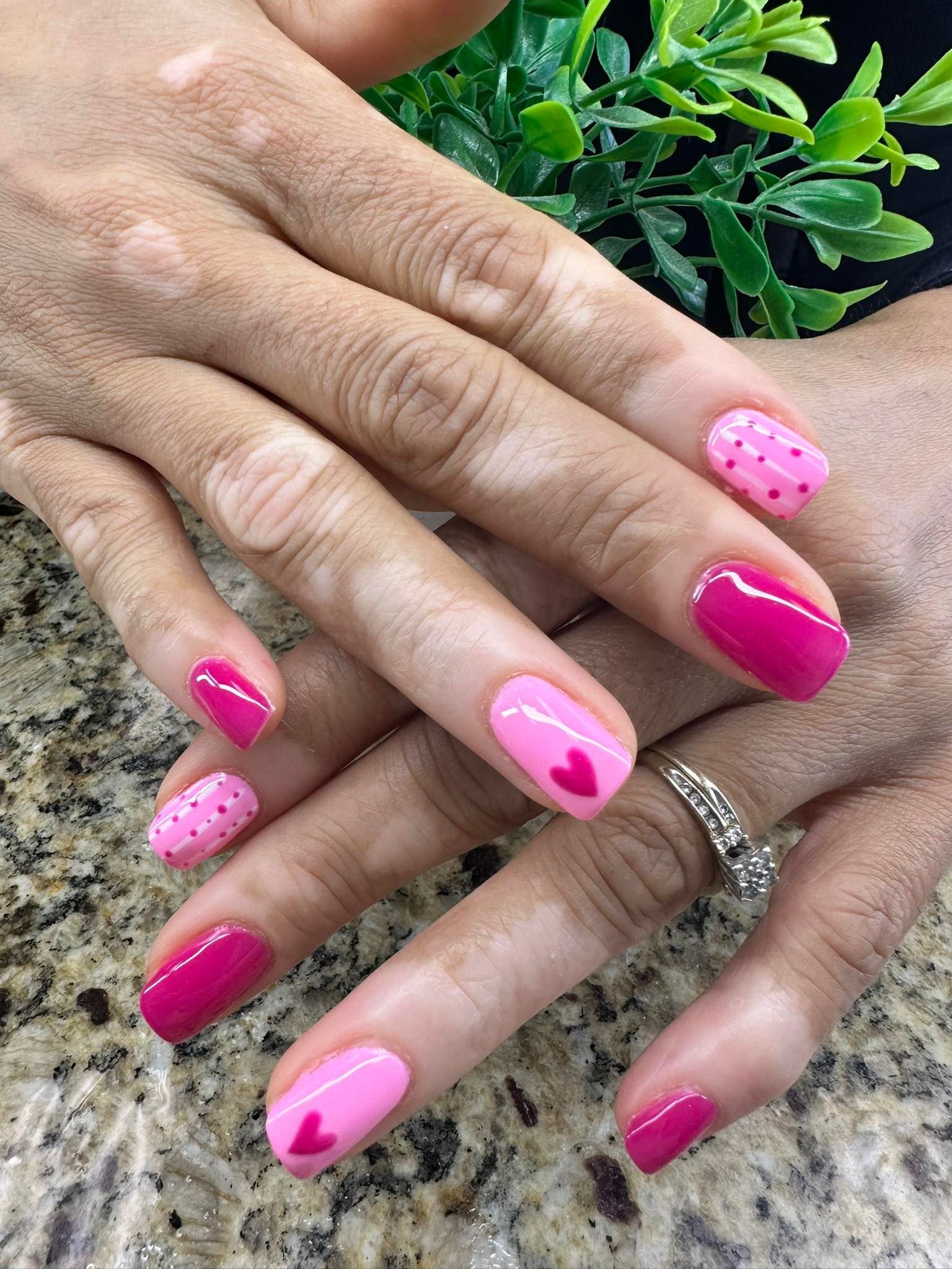 Hands with bright pink manicured nails resting on a granite countertop beside green leaves