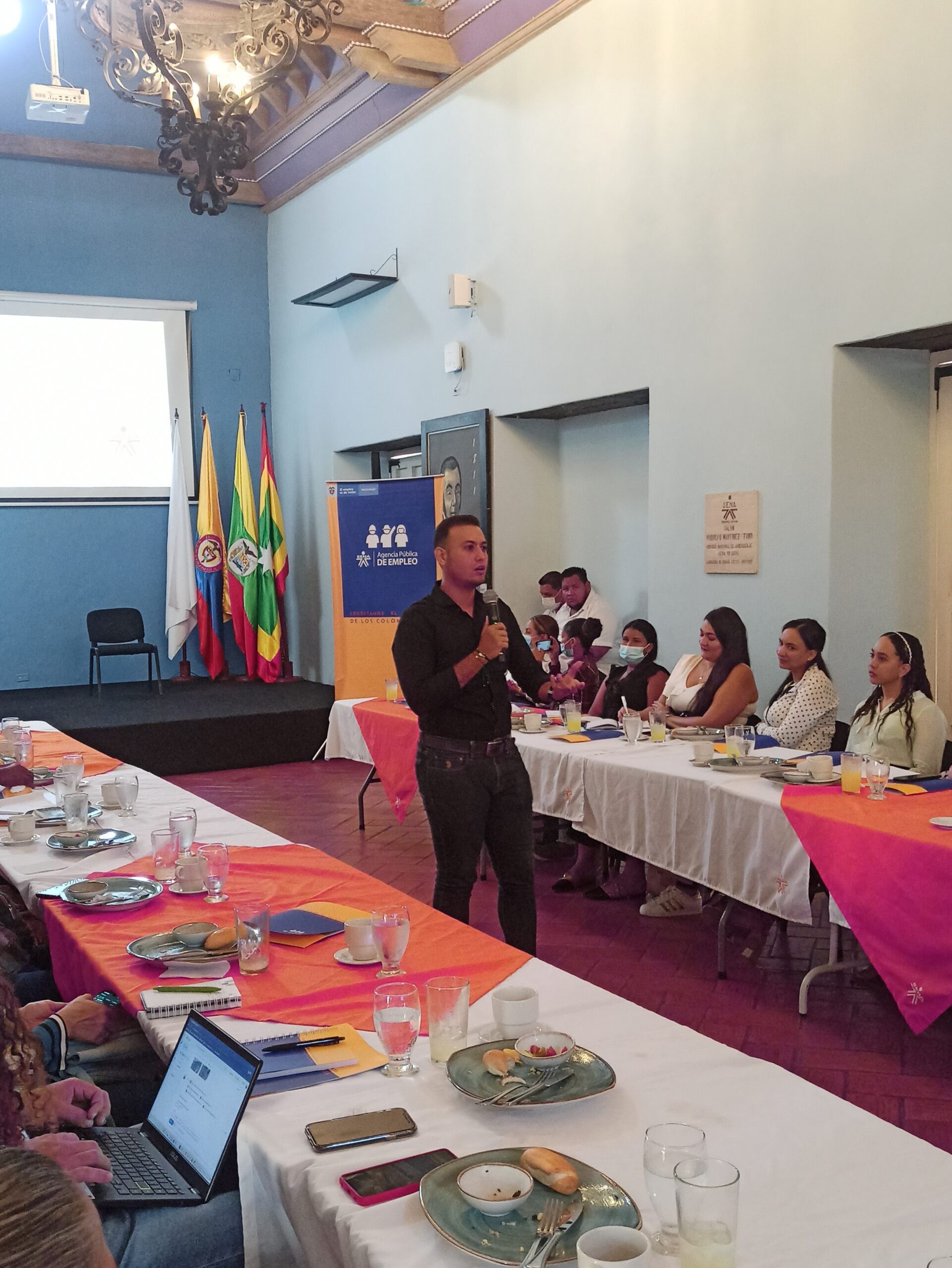 Un hombre está dando una presentación a un grupo de personas sentadas en mesas largas.