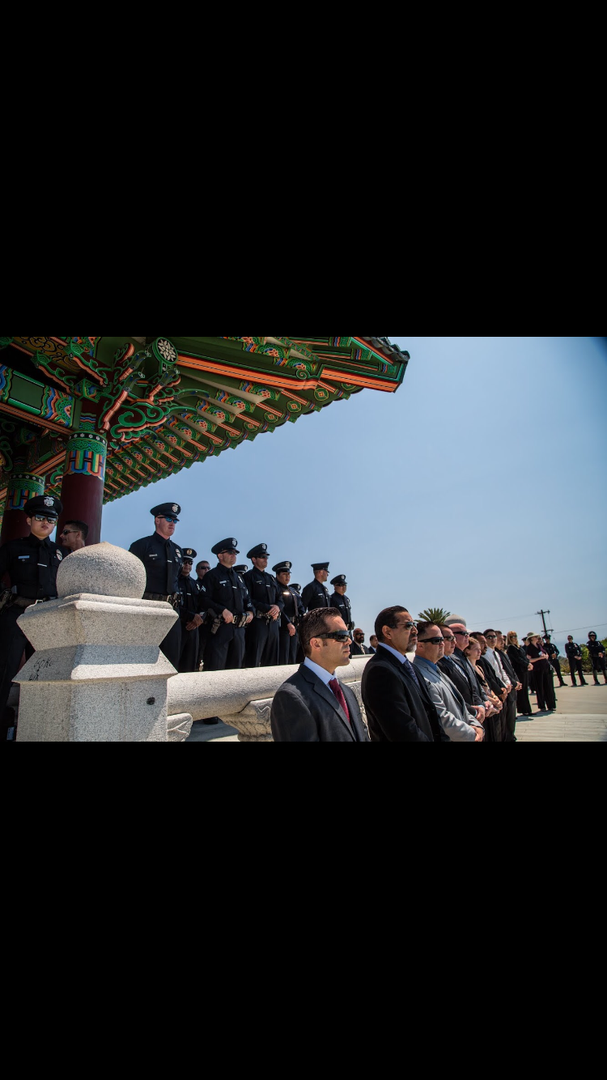 A line of police officers and men in suits standing in profile under a traditional Korean-style pavilion roof.
